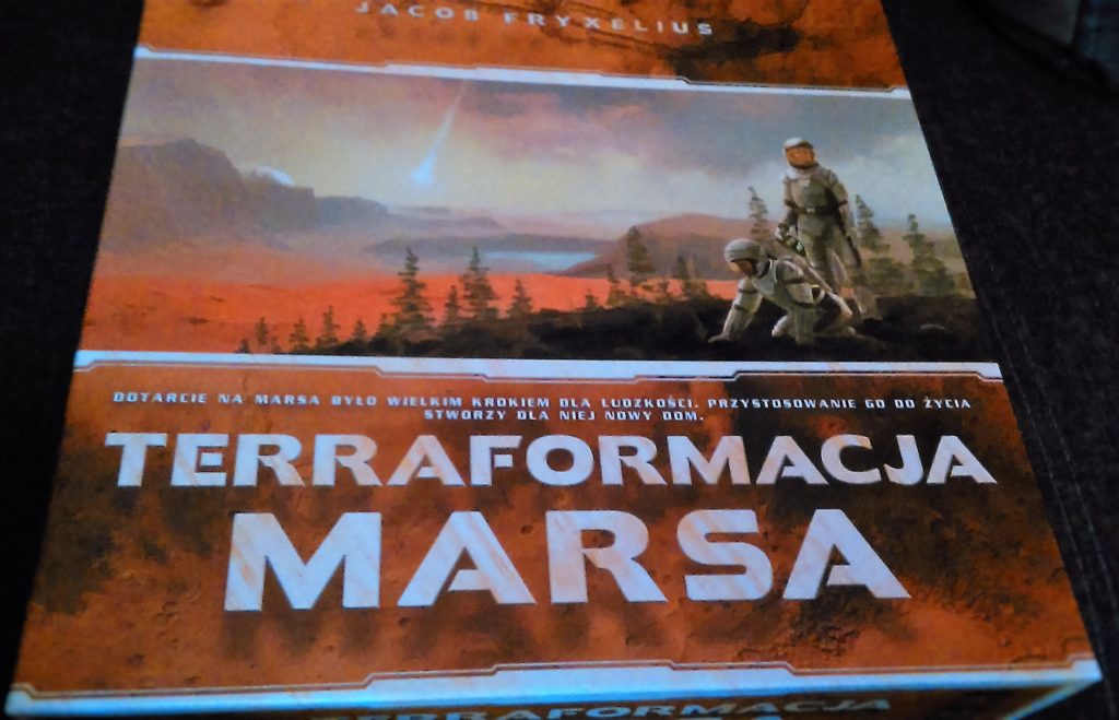 Wieczko pudełka do gry, z grającą pierwsze skrzypce grafiką przedstawiającą astronautów na Marsie.